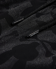 Close-up of Psycho Bunny's KIDS MALTA HYDROCHROMIC SWIM TRUNK - B0W321B2SW, showcasing a black textured fabric with a subtle camouflage pattern in shades of dark grey and black. Two parallel black cords with small text on one cord are partially visible, reminiscent of sleek swim trunks. The overall appearance is both modern and hydrochromic.