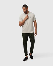 A man in a Psycho Bunny MENS CLASSIC PIQUE POLO SHIRT - B6K001B200, light beige, paired with dark green pants stands against a white background. He wears black slip-on shoes and has neat short hair. His left hand touches his neck while he gazes downwards, showcasing men's fashion.