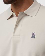 A close-up of someone wearing the Psycho Bunny MENS CLASSIC PIQUE POLO SHIRT - B6K001B200 in light beige, crafted from Pima cotton with two buttons undone. It features a small pink and white bunny with crossbones logo on the chest, representing timeless men's fashion.