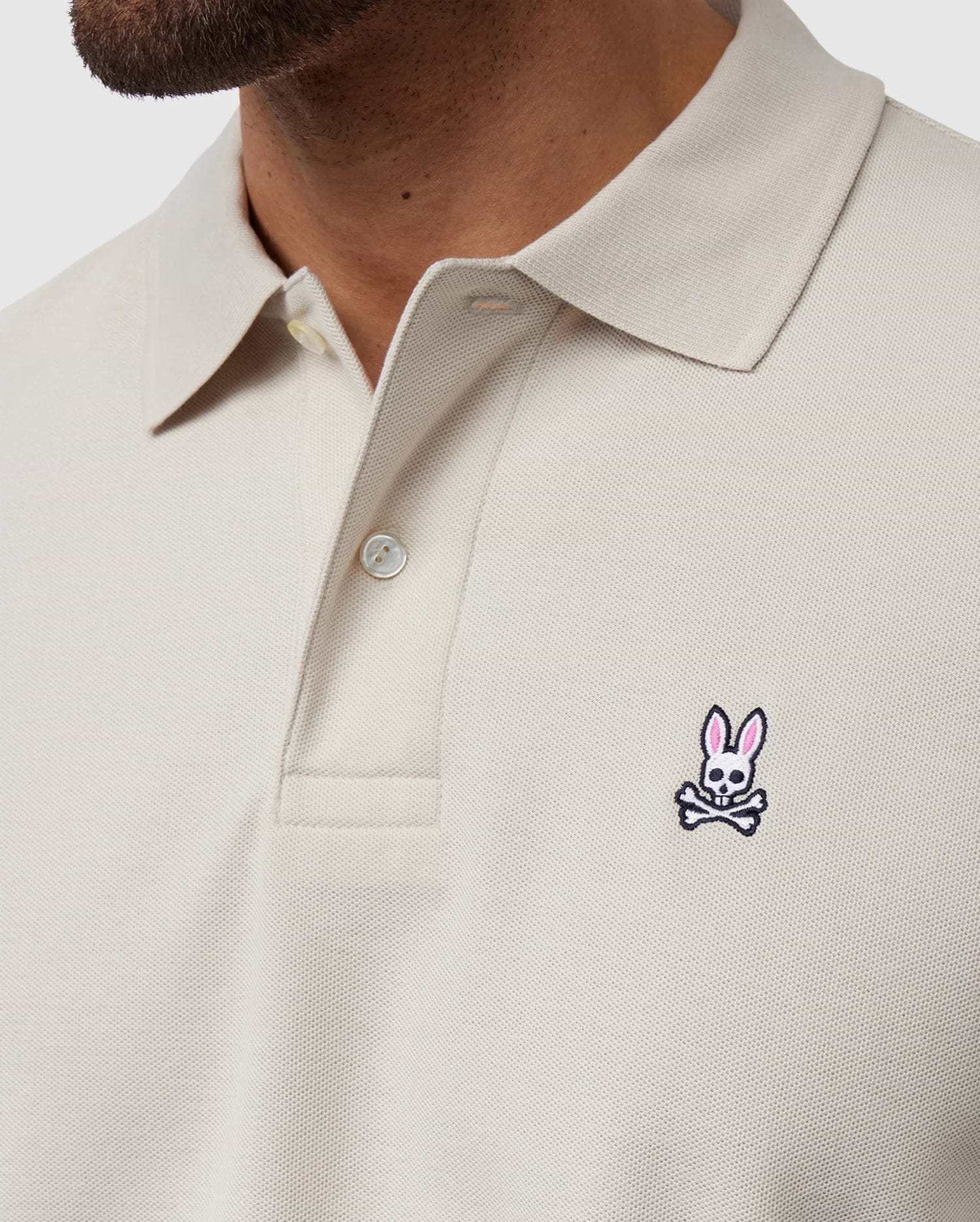 A close-up of someone wearing the Psycho Bunny MENS CLASSIC PIQUE POLO SHIRT - B6K001B200 in light beige, crafted from Pima cotton with two buttons undone. It features a small pink and white bunny with crossbones logo on the chest, representing timeless men's fashion.