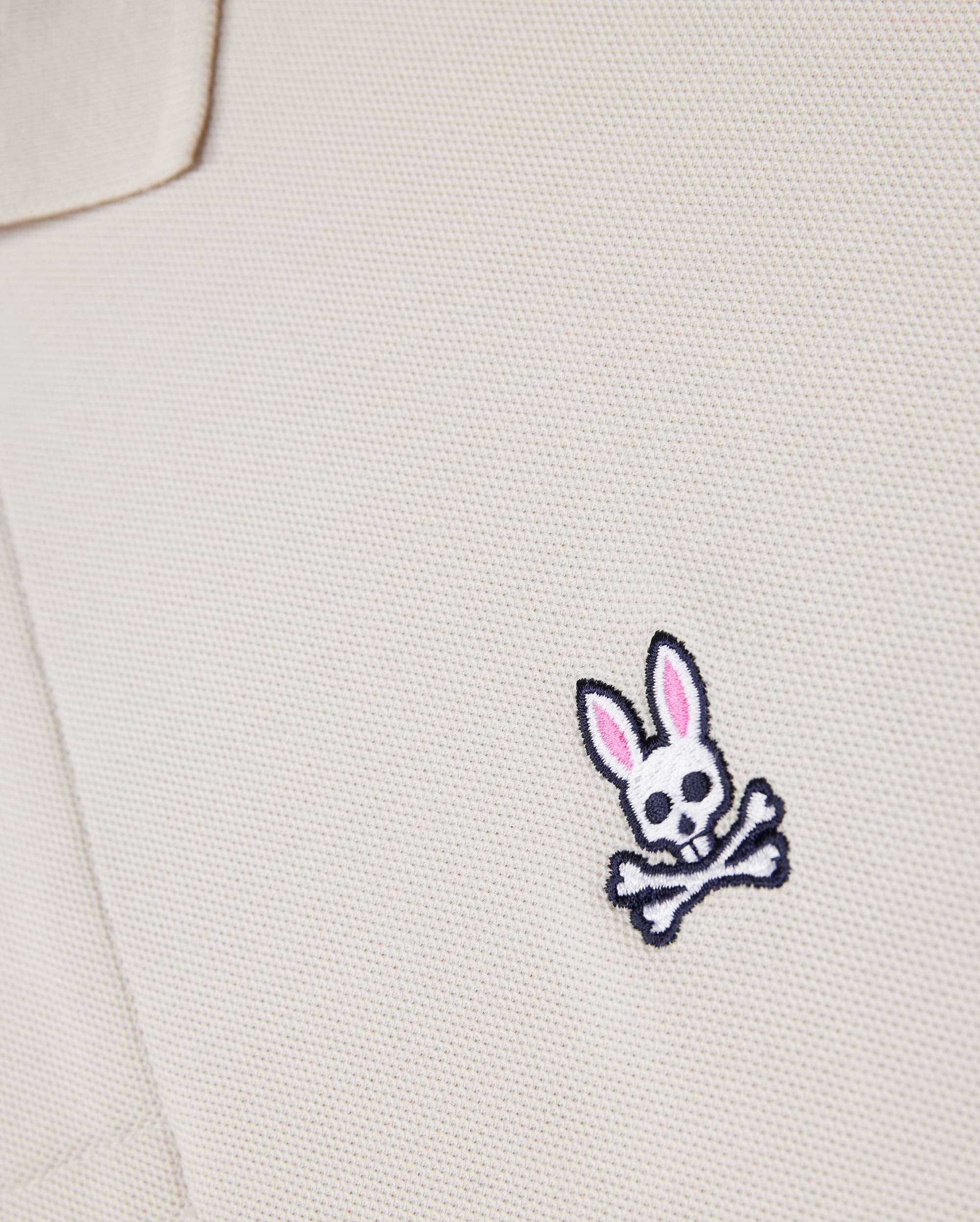 A close-up of the Psycho Bunny beige MENS CLASSIC PIQUE POLO SHIRT - B6K001B200, made from premium Pima cotton, showcases a small bunny skull emblem with pink ears and crossbones. It adds an edgy twist to men's fashion.