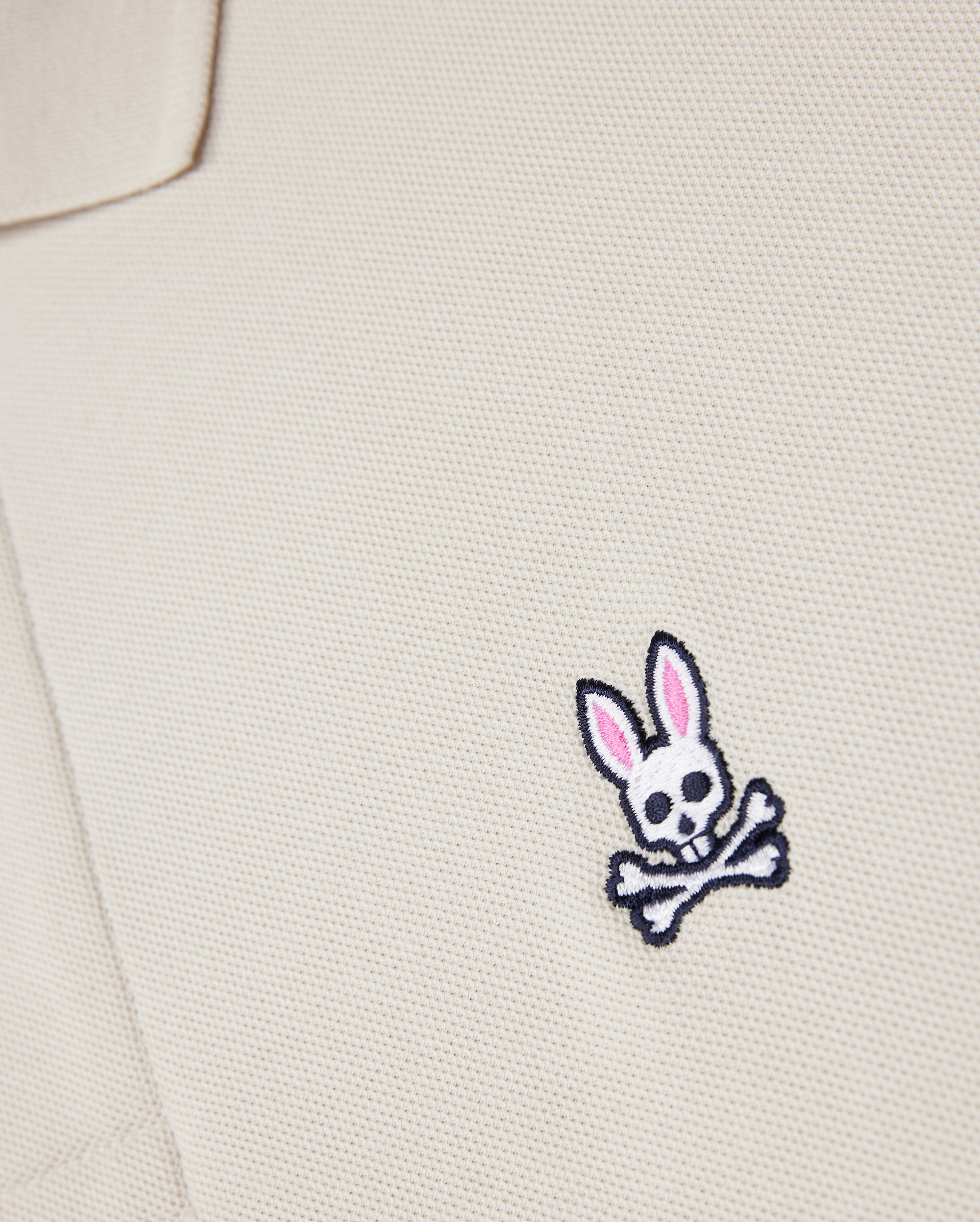 A close-up of the Psycho Bunny beige MENS CLASSIC PIQUE POLO SHIRT - B6K001B200, made from premium Pima cotton, showcases a small bunny skull emblem with pink ears and crossbones. It adds an edgy twist to men's fashion.