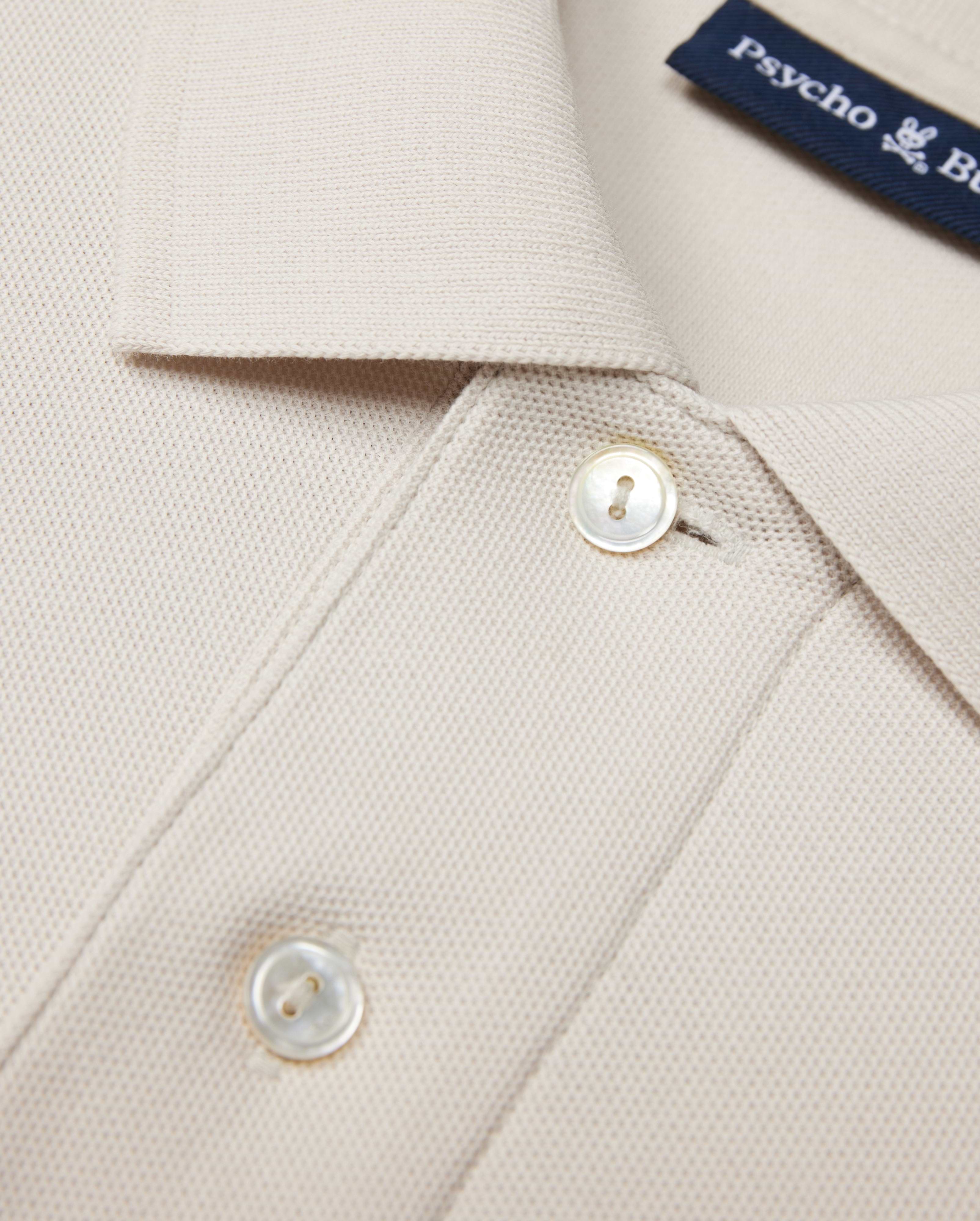 The Psycho Bunny MENS CLASSIC PIQUE POLO SHIRT - B6K001B200 in beige is crafted from luxurious Pima cotton with textured fabric. It features a collar, two white buttons, and a visible blue text label inside, embodying timeless men's fashion elegance.