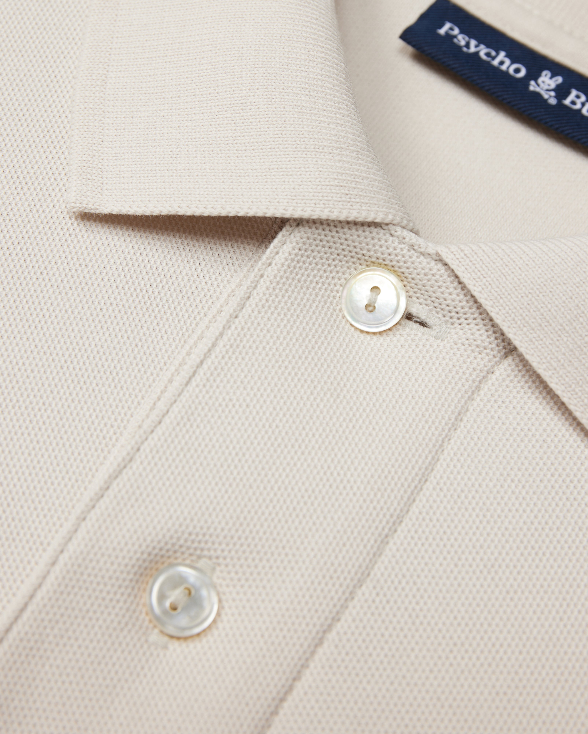 The Psycho Bunny MENS CLASSIC PIQUE POLO SHIRT - B6K001B200 in beige is crafted from luxurious Pima cotton with textured fabric. It features a collar, two white buttons, and a visible blue text label inside, embodying timeless men's fashion elegance.