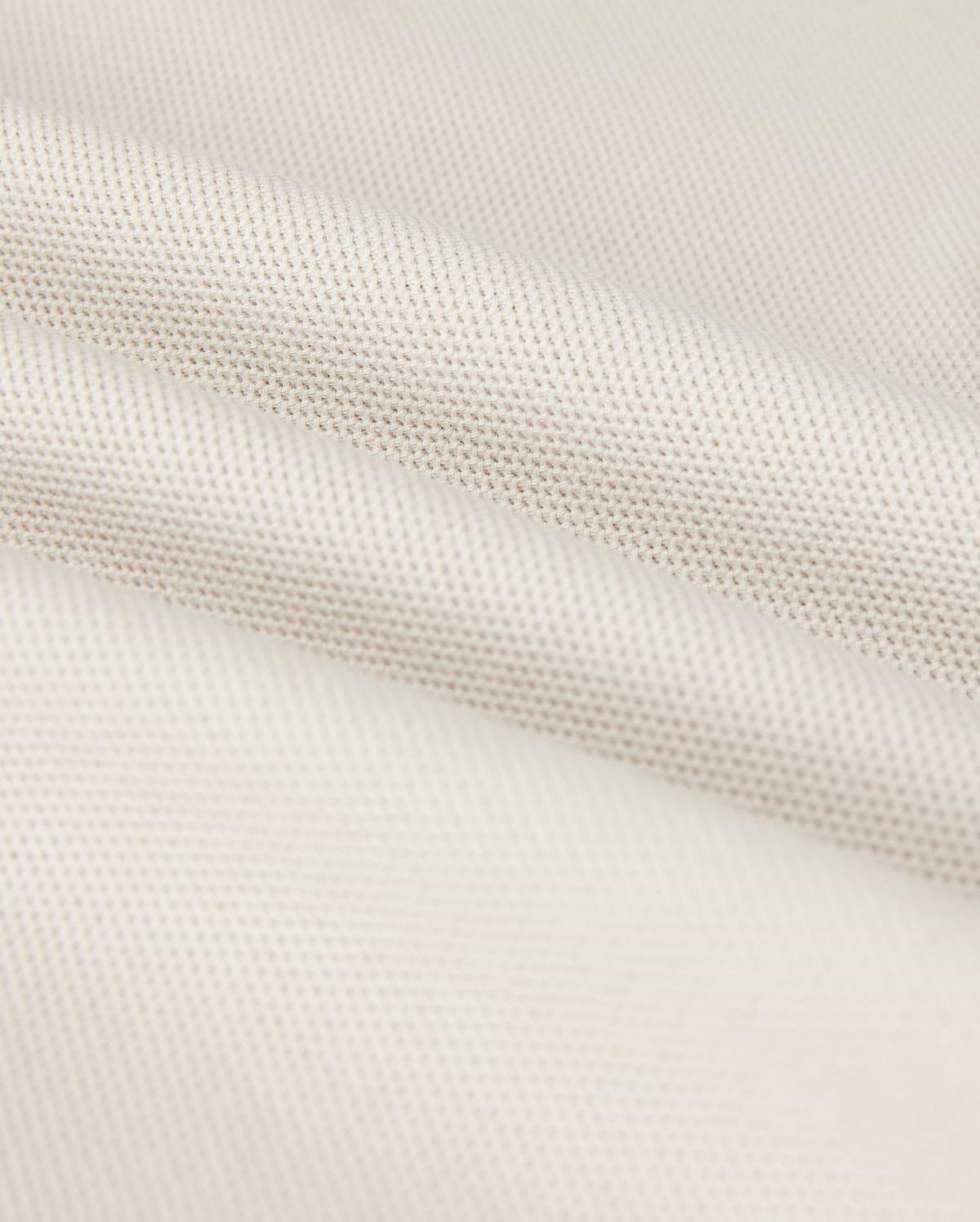 A close-up shows the Psycho Bunny MENS CLASSIC PIQUE POLO SHIRT - B6K001B200, with a textured weave pattern. The soft white fabric resembles fine Pima cotton, with subtle shadowing in the folds that exude timeless men's fashion.