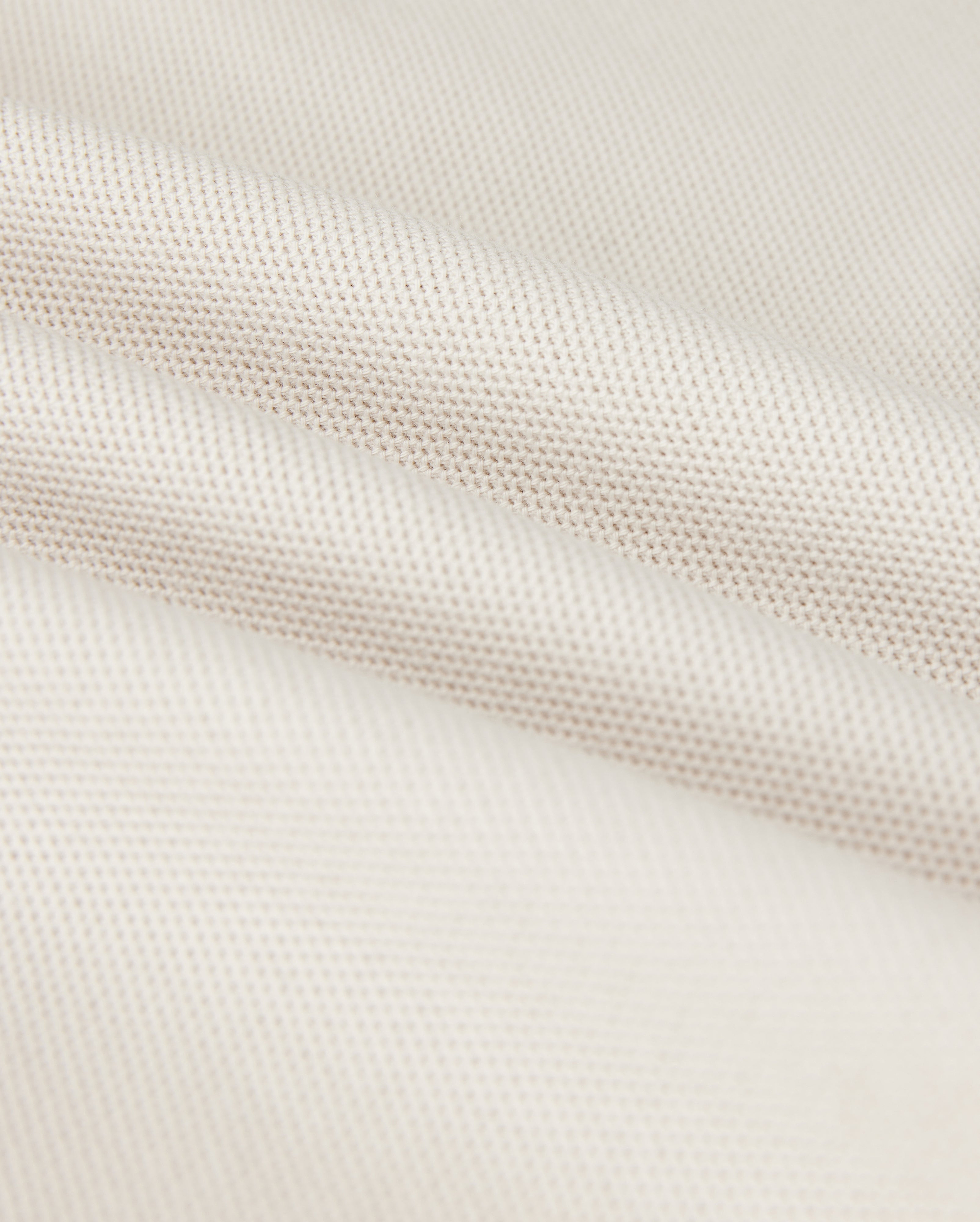 A close-up shows the Psycho Bunny MENS CLASSIC PIQUE POLO SHIRT - B6K001B200, with a textured weave pattern. The soft white fabric resembles fine Pima cotton, with subtle shadowing in the folds that exude timeless men's fashion.