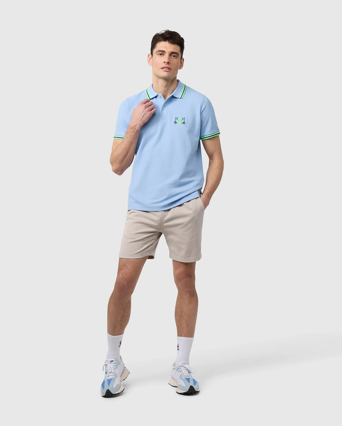 A person dressed in a light blue polo shirt with green accents, paired with the MENS WILLIS STRETCH TENCEL SHORT - B6R239Y1WB by Psycho Bunny, along with white socks and sneakers, stands against a plain background. The shirt is adorned with a small green logo on the chest.