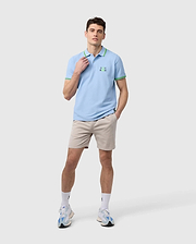 A person dressed in a light blue polo shirt with green accents, paired with the MENS WILLIS STRETCH TENCEL SHORT - B6R239Y1WB by Psycho Bunny, along with white socks and sneakers, stands against a plain background. The shirt is adorned with a small green logo on the chest.
