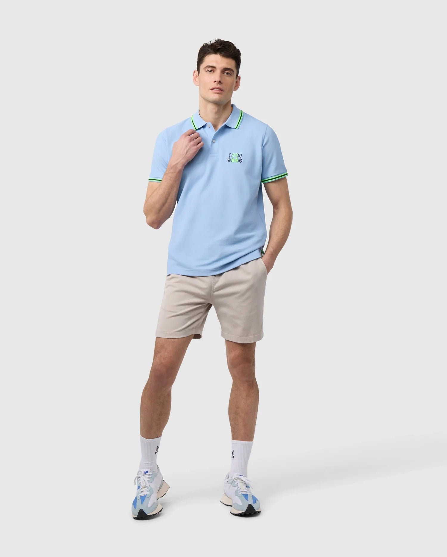 A person dressed in a light blue polo shirt with green accents, paired with the MENS WILLIS STRETCH TENCEL SHORT - B6R239Y1WB by Psycho Bunny, along with white socks and sneakers, stands against a plain background. The shirt is adorned with a small green logo on the chest.