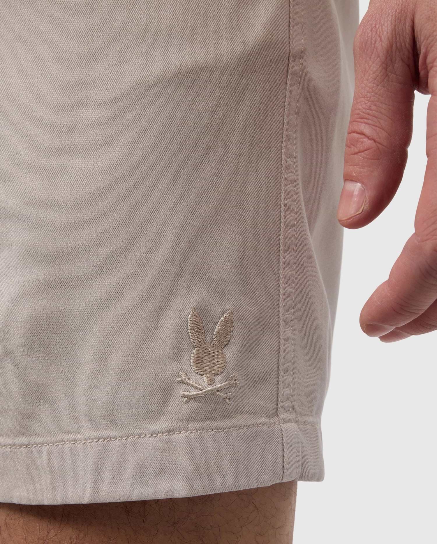 Someone sporting the eco-friendly MENS WILLIS STRETCH TENCEL SHORT - B6R239Y1WB by Psycho Bunny, featuring an embroidered bunny and crossbones logo on the side. The right hand is partially visible, showcasing the comfort and style of these sustainable shorts.