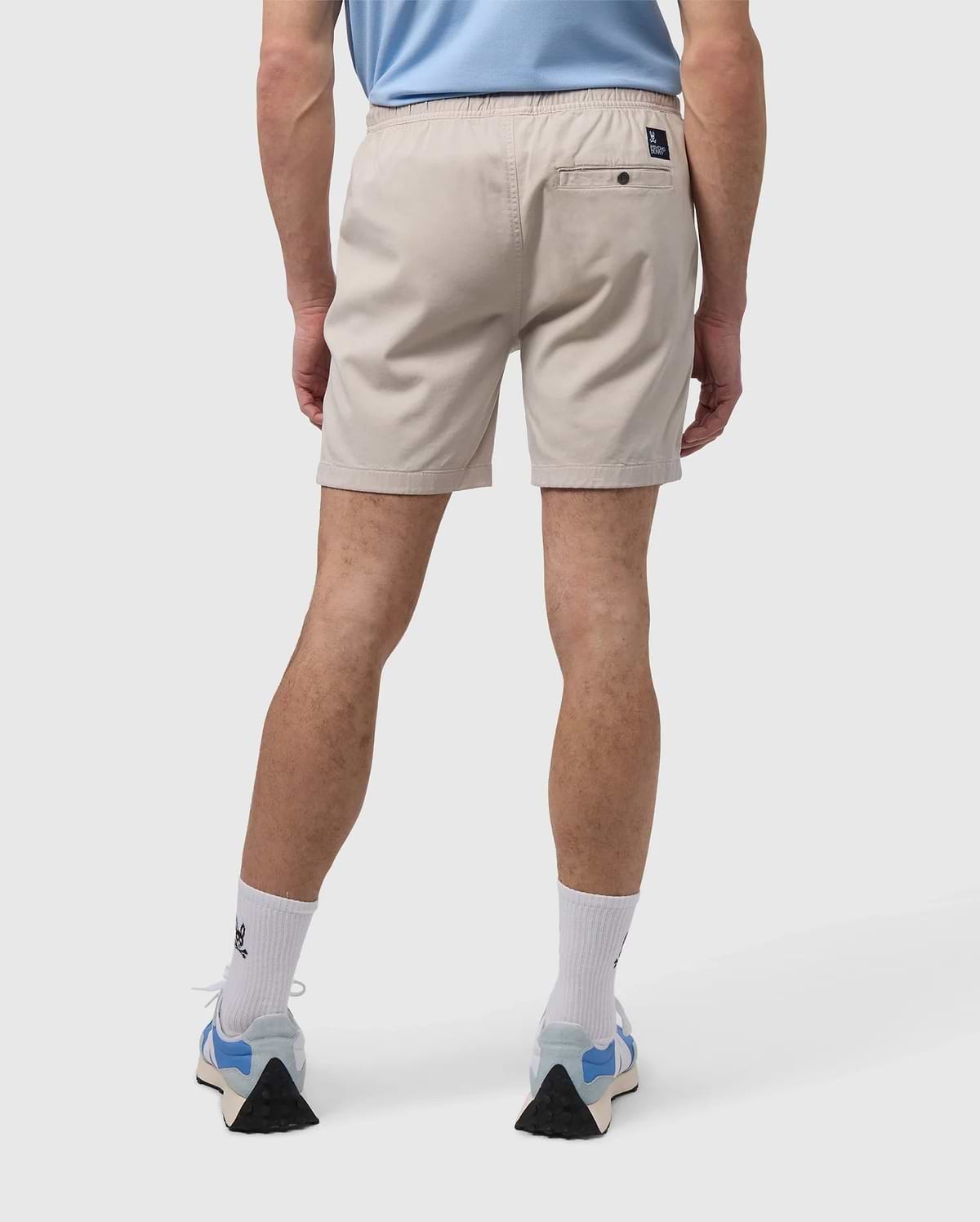 A person is wearing a light blue shirt, Psycho Bunny's MENS WILLIS STRETCH TENCEL SHORT in beige, white socks, and blue sneakers while standing against a plain background, shown from the back.