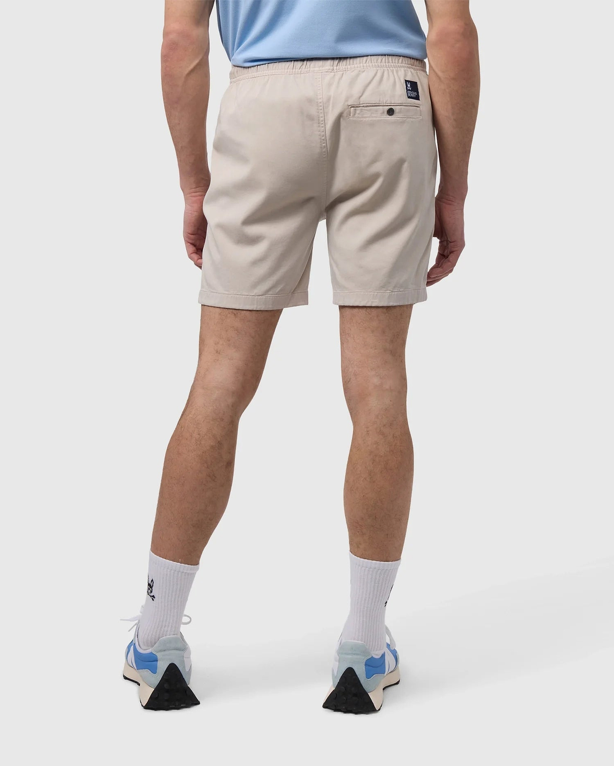 A person is wearing a light blue shirt, Psycho Bunny's MENS WILLIS STRETCH TENCEL SHORT in beige, white socks, and blue sneakers while standing against a plain background, shown from the back.