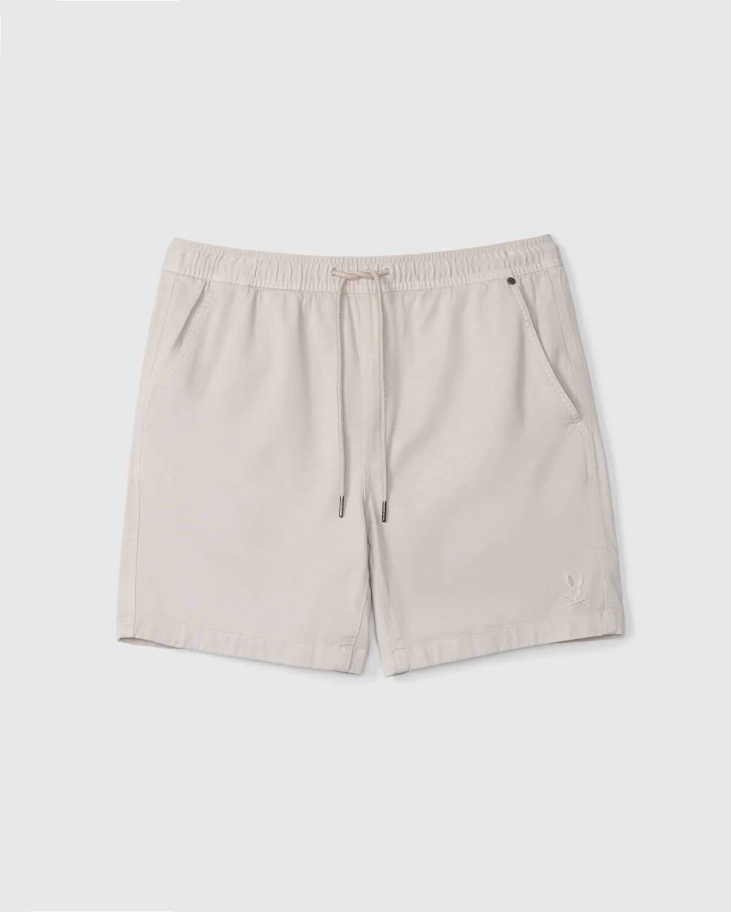 A pair of beige Men's Willis Stretch TENCEL™ shorts from Psycho Bunny, featuring an elastic waistband with a drawstring, two front pockets, and a small embroidered logo on the lower left. The lightweight fabric makes them ideal for casual wear.