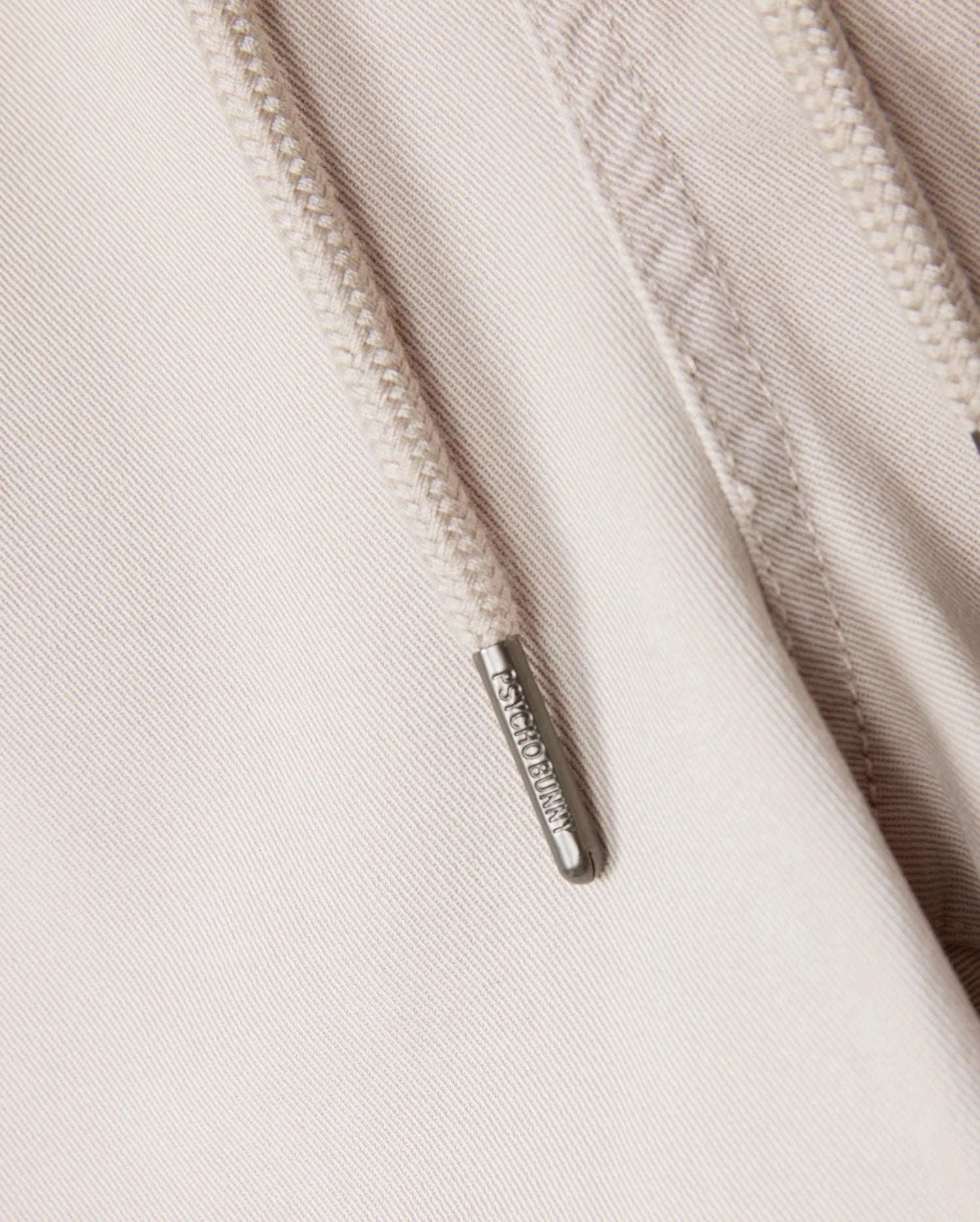 Close-up of a beige hoodie showcasing a metal zipper pull engraved with "ARMANI EXCHANGE." The visible fabric texture and detailed stitching around the zipper make it an ideal pairing with the Psycho Bunny MENS WILLIS STRETCH TENCEL SHORTS or elasticated waist trousers.