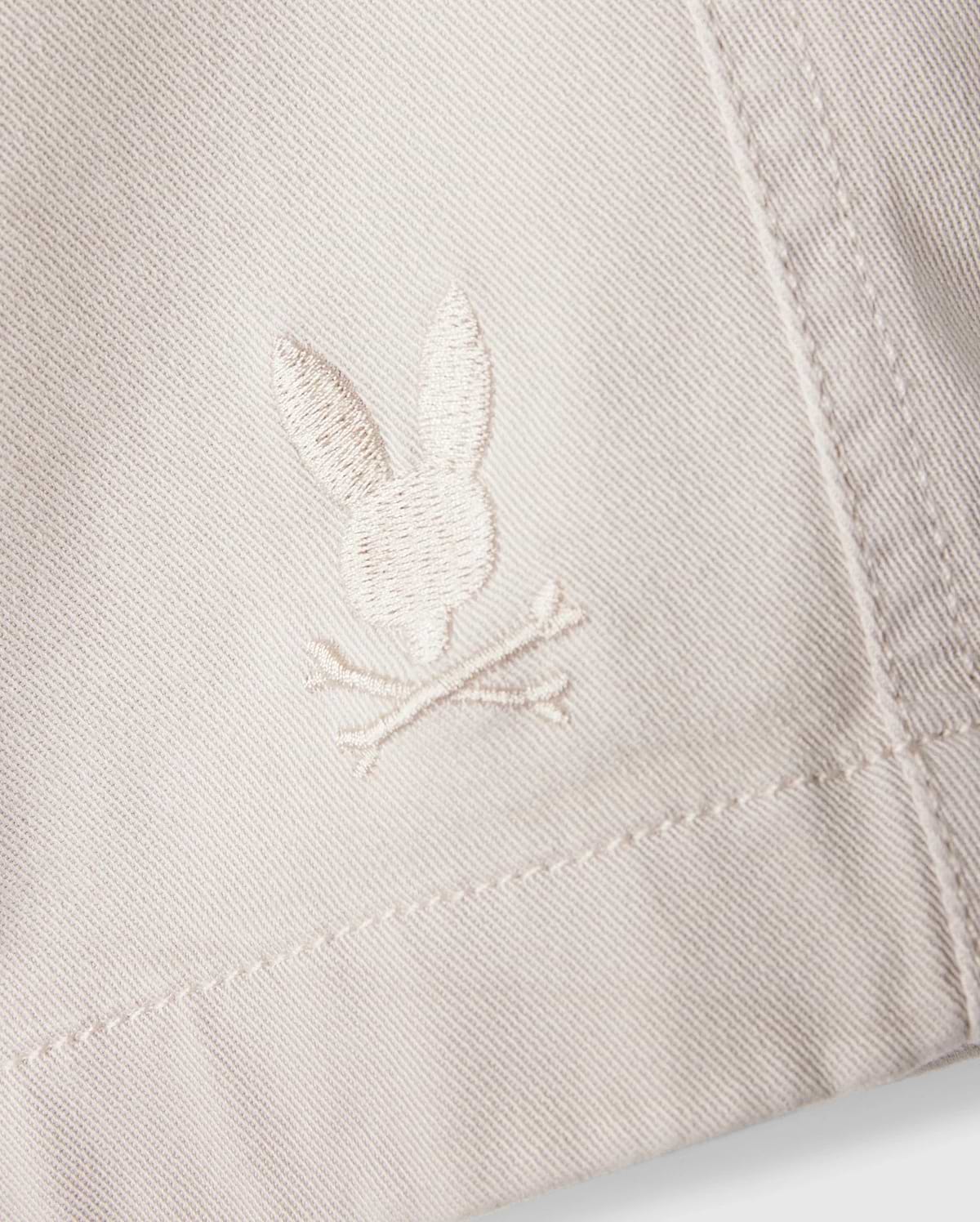 A close-up of the white fabric on the Psycho Bunny MENS WILLIS STRETCH TENCEL SHORT - B6R239Y1WB reveals an embroidered logo featuring a rabbit head and crossbones. The stitching seamlessly blends with the fabric's hue, while the texture accentuates the embroidery's details, providing an eco-friendly twist to stylish comfort.
