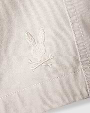 A close-up of the white fabric on the Psycho Bunny MENS WILLIS STRETCH TENCEL SHORT - B6R239Y1WB reveals an embroidered logo featuring a rabbit head and crossbones. The stitching seamlessly blends with the fabric's hue, while the texture accentuates the embroidery's details, providing an eco-friendly twist to stylish comfort.