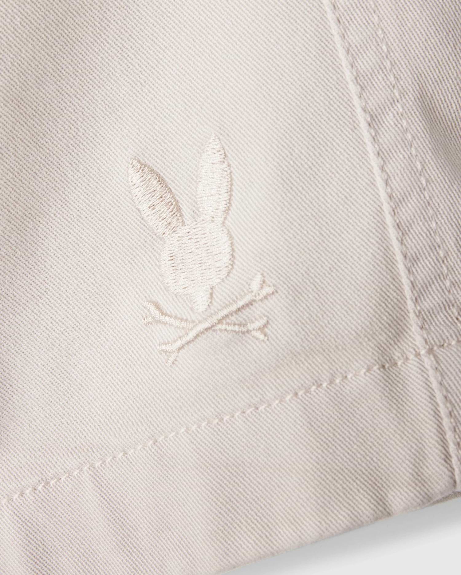 A close-up of the white fabric on the Psycho Bunny MENS WILLIS STRETCH TENCEL SHORT - B6R239Y1WB reveals an embroidered logo featuring a rabbit head and crossbones. The stitching seamlessly blends with the fabric's hue, while the texture accentuates the embroidery's details, providing an eco-friendly twist to stylish comfort.