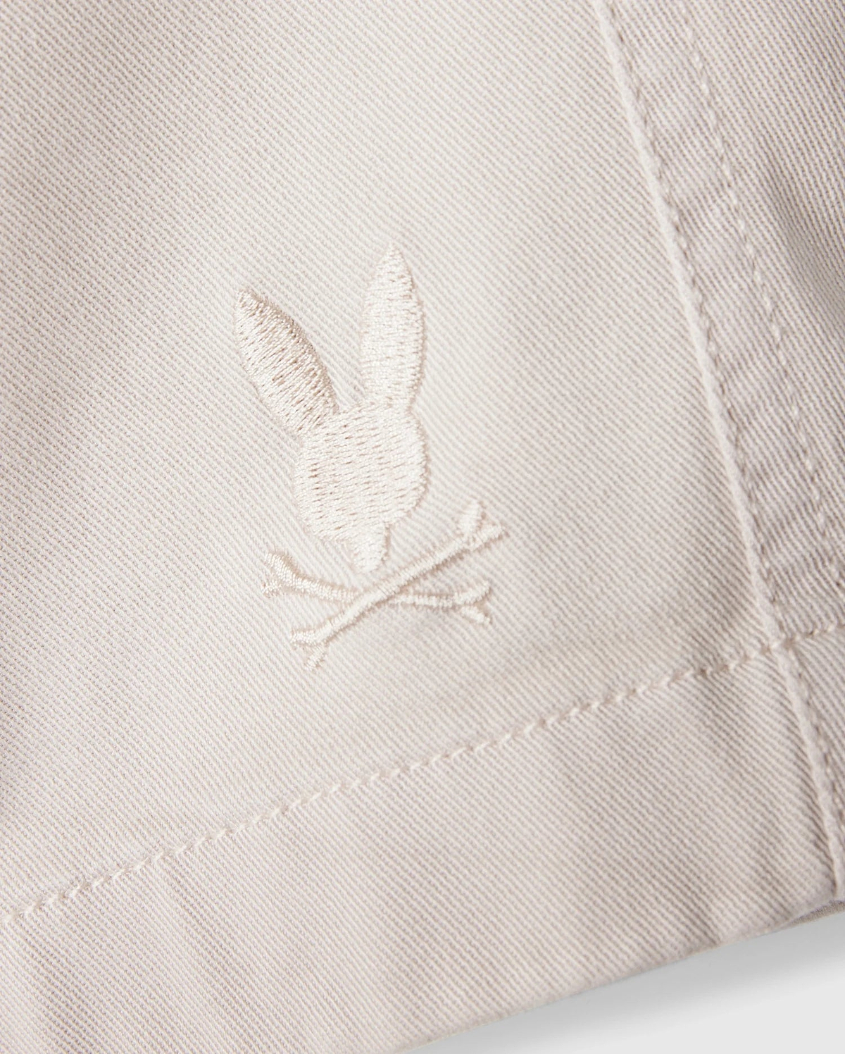 A close-up of the white fabric on the Psycho Bunny MENS WILLIS STRETCH TENCEL SHORT - B6R239Y1WB reveals an embroidered logo featuring a rabbit head and crossbones. The stitching seamlessly blends with the fabric's hue, while the texture accentuates the embroidery's details, providing an eco-friendly twist to stylish comfort.