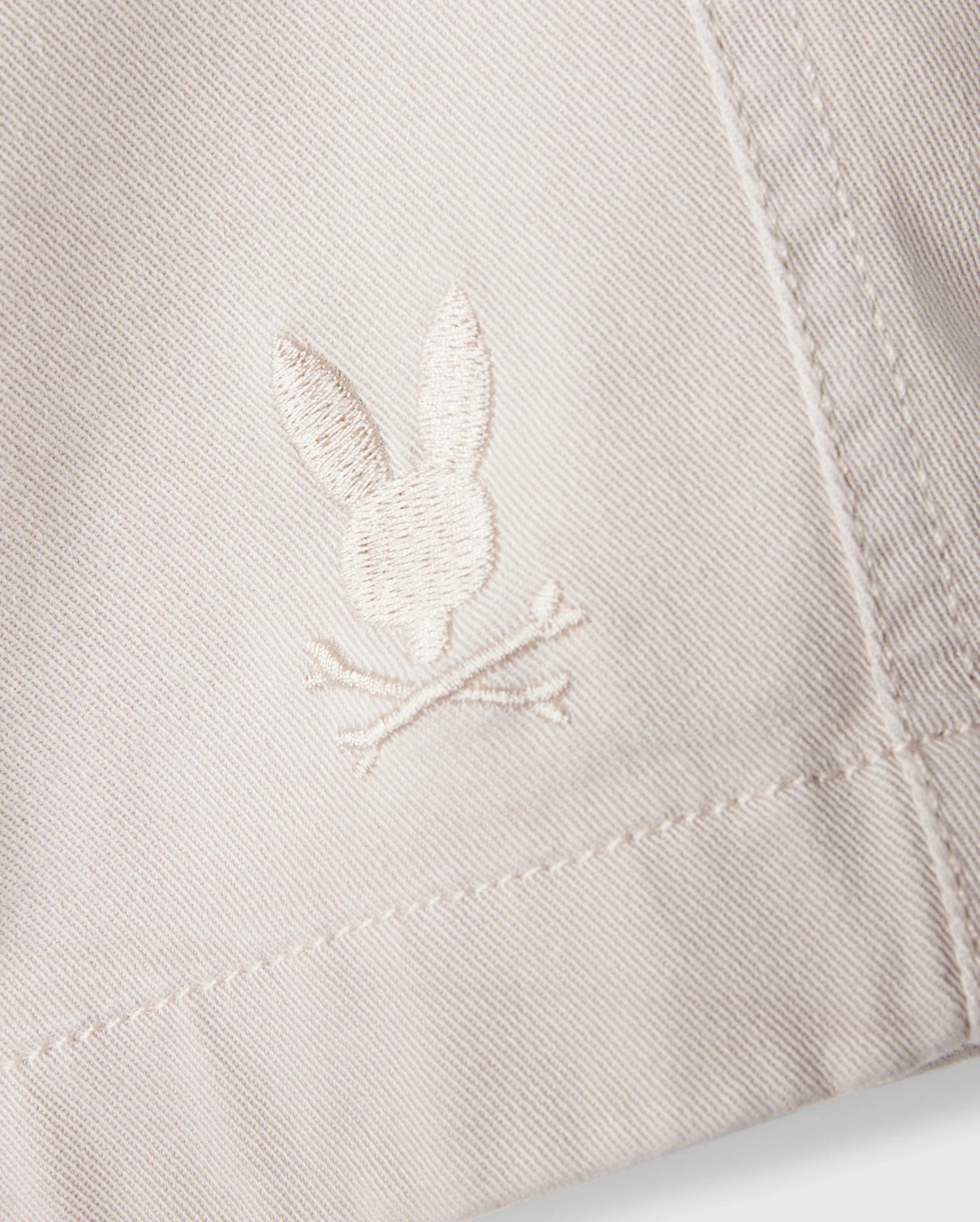 A close-up of the white fabric on the Psycho Bunny MENS WILLIS STRETCH TENCEL SHORT - B6R239Y1WB reveals an embroidered logo featuring a rabbit head and crossbones. The stitching seamlessly blends with the fabric's hue, while the texture accentuates the embroidery's details, providing an eco-friendly twist to stylish comfort.