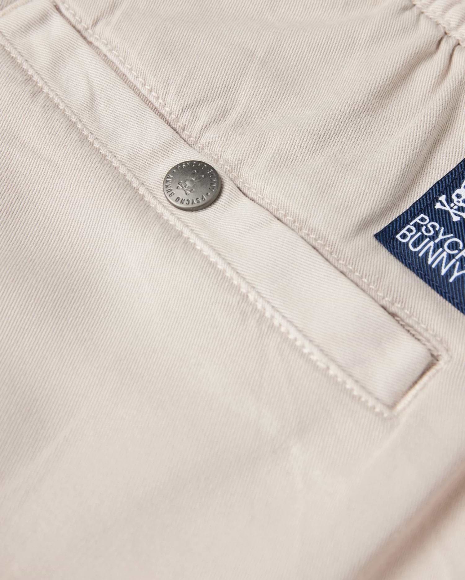 Close-up of the beige fabric from the Men's Willis Stretch TENCEL Shorts (B6R239Y1WB), showcasing a silver button embossed with "Psycho Bunny." A blue label featuring a white logo and text is sewn next to the pocket, completing these stylish eco-friendly men's shorts.