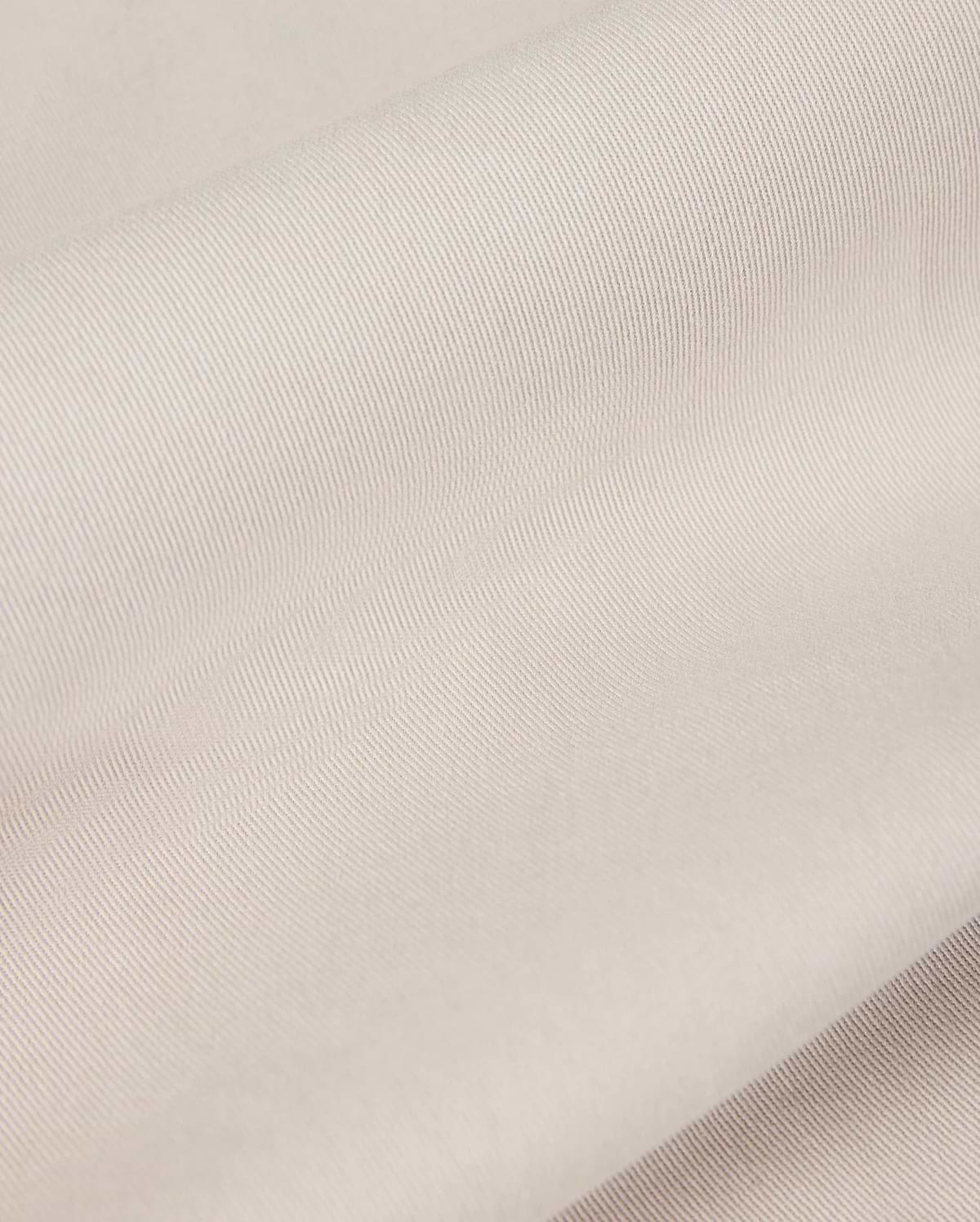 Close-up of the light beige fabric from the Psycho Bunny MENS WILLIS STRETCH TENCEL SHORT - B6R239Y1WB, featuring a smooth, slightly ribbed texture that is perfect for men's trousers. The material looks soft and supple, gently folded in overlapping layers to highlight its subtle sheen and fine weave pattern.