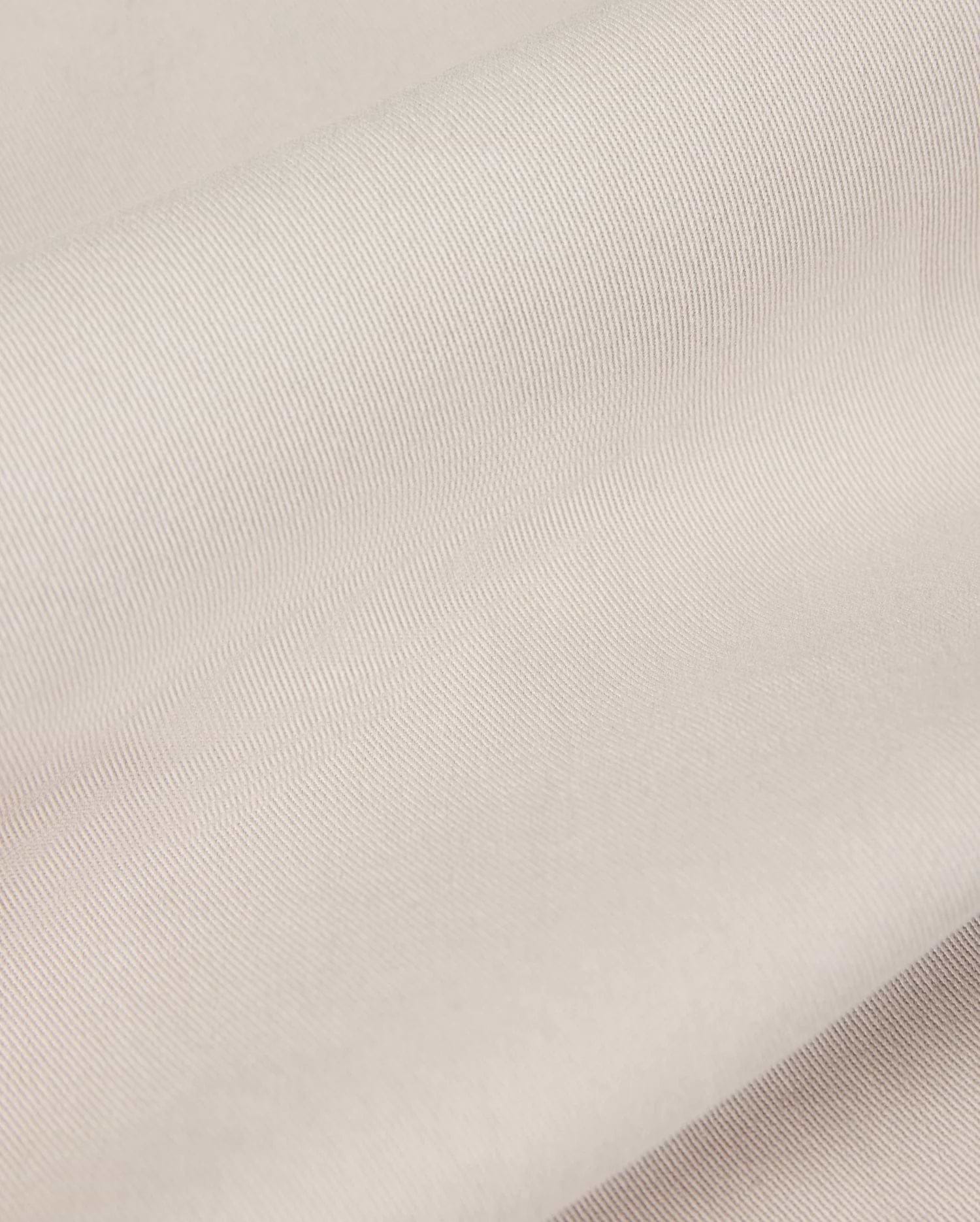 Close-up of the light beige fabric from the Psycho Bunny MENS WILLIS STRETCH TENCEL SHORT - B6R239Y1WB, featuring a smooth, slightly ribbed texture that is perfect for men's trousers. The material looks soft and supple, gently folded in overlapping layers to highlight its subtle sheen and fine weave pattern.