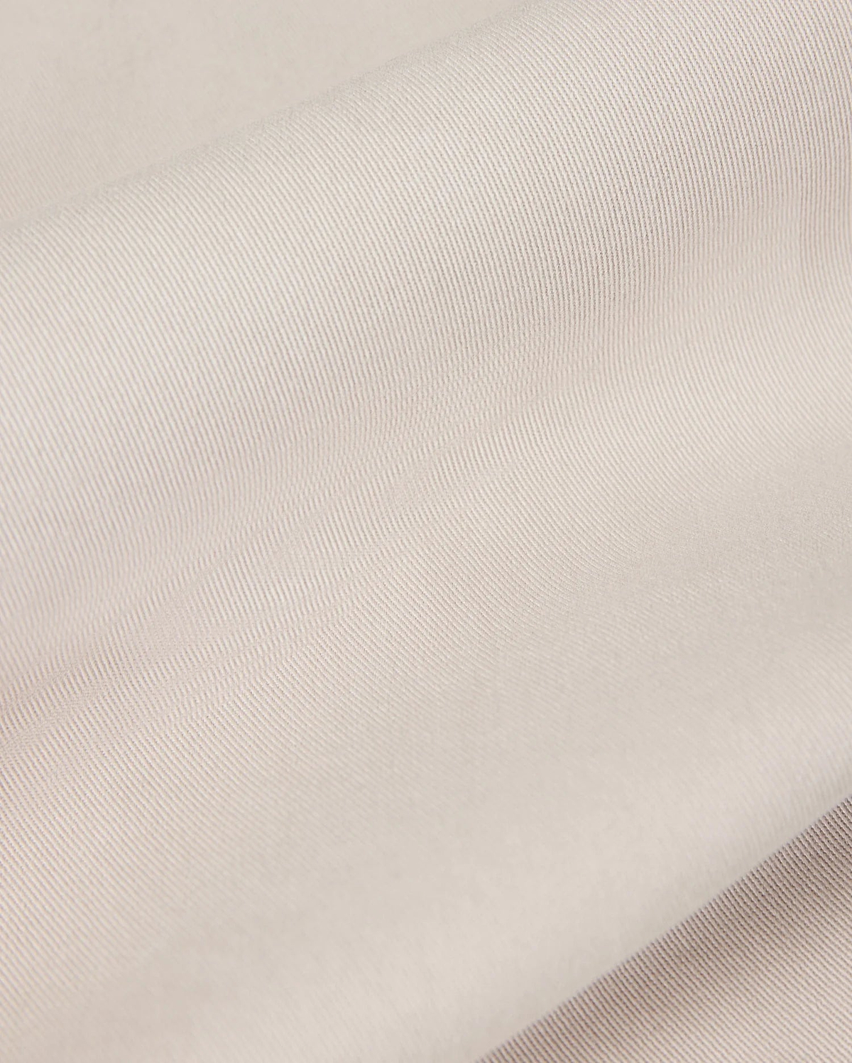 Close-up of the light beige fabric from the Psycho Bunny MENS WILLIS STRETCH TENCEL SHORT - B6R239Y1WB, featuring a smooth, slightly ribbed texture that is perfect for men's trousers. The material looks soft and supple, gently folded in overlapping layers to highlight its subtle sheen and fine weave pattern.