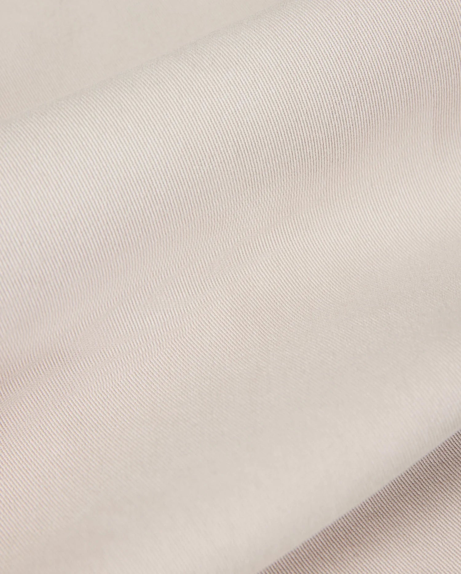 Close-up of the light beige fabric from the Psycho Bunny MENS WILLIS STRETCH TENCEL SHORT - B6R239Y1WB, featuring a smooth, slightly ribbed texture that is perfect for men's trousers. The material looks soft and supple, gently folded in overlapping layers to highlight its subtle sheen and fine weave pattern.