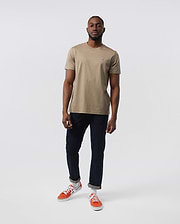 A man stands against a plain, white background wearing the Psycho Bunny MENS CLASSIC CREW NECK TEE - B6U014CRPC in beige, crafted from 100% Pima cotton. Paired with dark blue jeans and orange sneakers, his look forms the basis for any man's wardrobe. He has a short beard and close-cropped hair, with his right hand partially in his pocket.