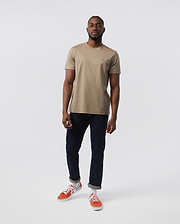 A man stands against a plain, white background wearing the Psycho Bunny MENS CLASSIC CREW NECK TEE - B6U014CRPC in beige, crafted from 100% Pima cotton. Paired with dark blue jeans and orange sneakers, his look forms the basis for any man's wardrobe. He has a short beard and close-cropped hair, with his right hand partially in his pocket.