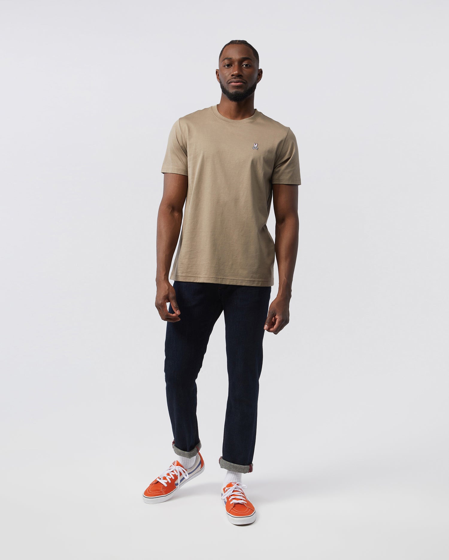A man stands against a plain, white background wearing the Psycho Bunny MENS CLASSIC CREW NECK TEE - B6U014CRPC in beige, crafted from 100% Pima cotton. Paired with dark blue jeans and orange sneakers, his look forms the basis for any man's wardrobe. He has a short beard and close-cropped hair, with his right hand partially in his pocket.