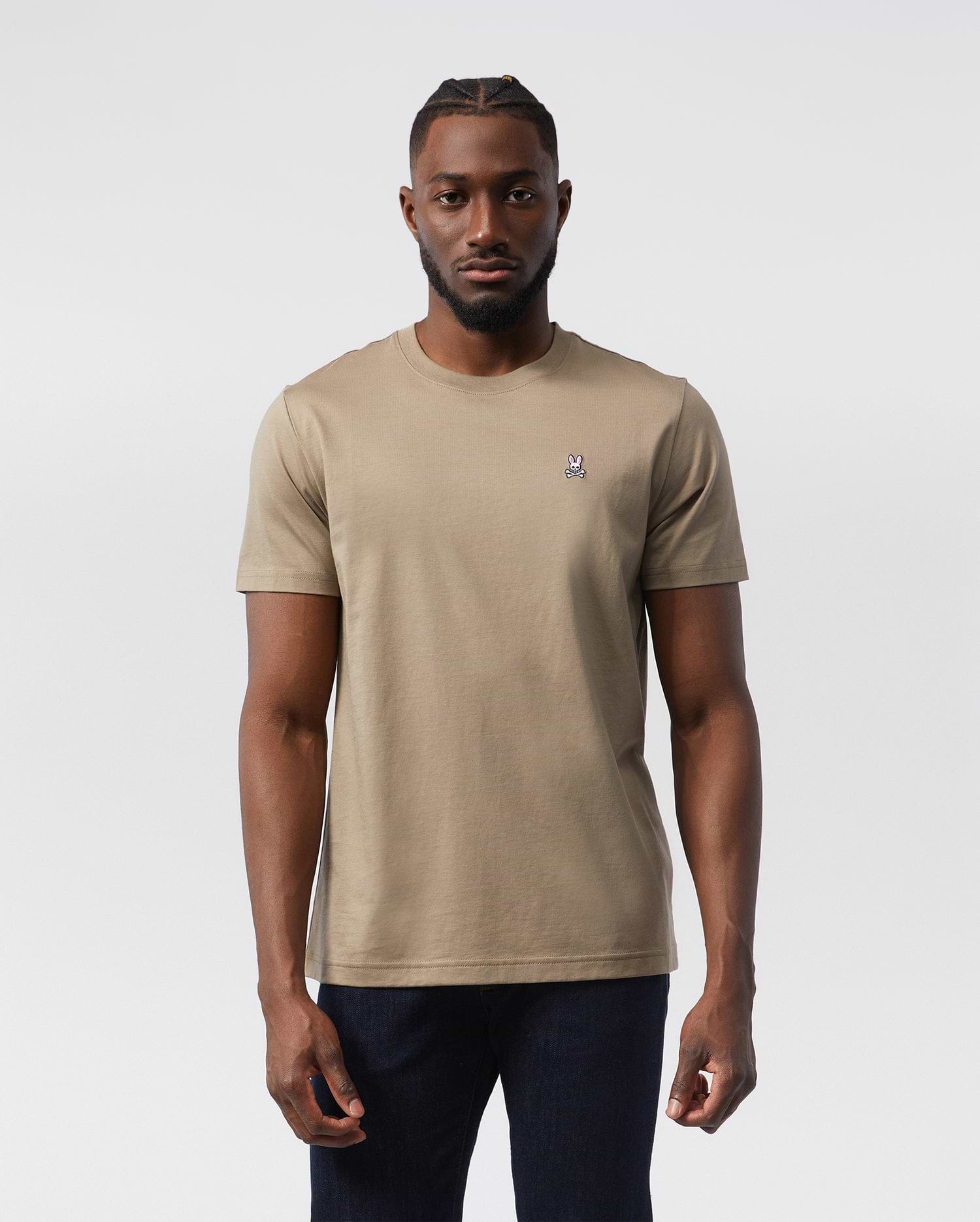 A man stands facing forward wearing a beige, 100% Pima cotton Psycho Bunny MENS CLASSIC CREW NECK TEE - B6U014CRPC with a small embroidered logo on the left chest. Sporting short hair with a styled pattern on top and a beard, he pairs his classic crew neck tee with dark jeans. The background is plain white.