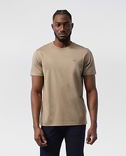 A man stands facing forward wearing a beige, 100% Pima cotton Psycho Bunny MENS CLASSIC CREW NECK TEE - B6U014CRPC with a small embroidered logo on the left chest. Sporting short hair with a styled pattern on top and a beard, he pairs his classic crew neck tee with dark jeans. The background is plain white.