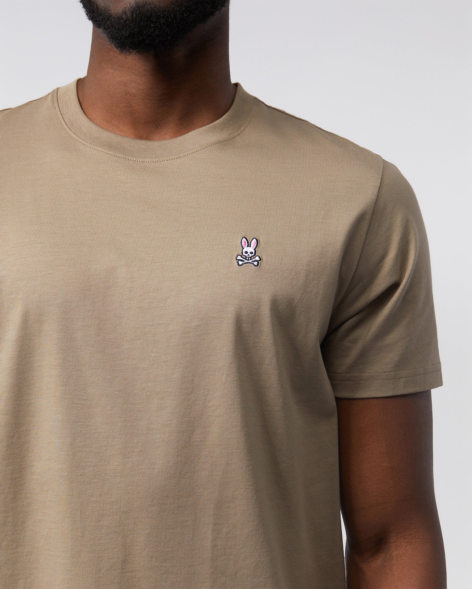 A person wearing a Psycho Bunny Men's Classic Crew Neck Tee - B6U014CRPC made from 100% Pima cotton. The beige t-shirt features a small embroidered logo of a pink bunny head with crossbones beneath it on the left side of the chest. The background is plain and white, with the person's lower face and neck visible.