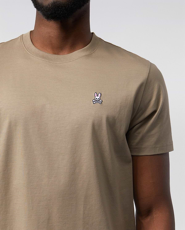 A person wearing a Psycho Bunny Men's Classic Crew Neck Tee - B6U014CRPC made from 100% Pima cotton. The beige t-shirt features a small embroidered logo of a pink bunny head with crossbones beneath it on the left side of the chest. The background is plain and white, with the person's lower face and neck visible.