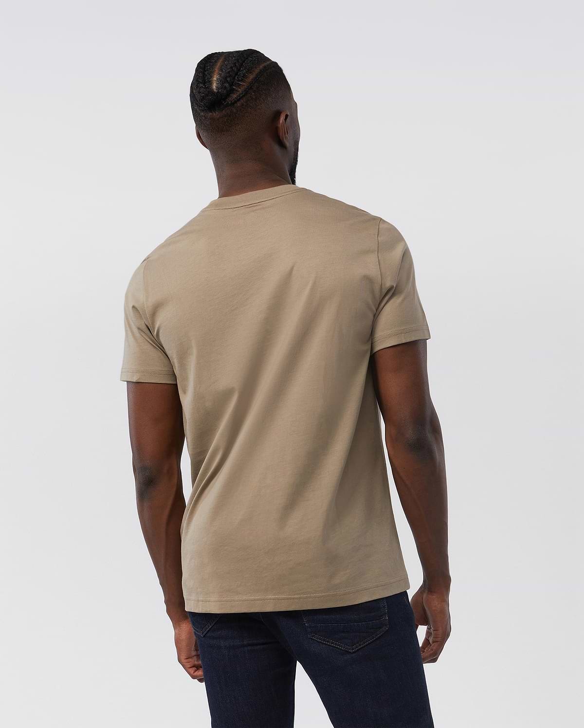 A person with cornrowed hair is seen from the back, wearing the Psycho Bunny MENS CLASSIC CREW NECK TEE - B6U014CRPC made of 100% Pima cotton and dark jeans. The light beige short-sleeve shirt serves as the basis for any man's wardrobe, standing out against the plain white background and highlighting the individual's casual attire and hairstyle.