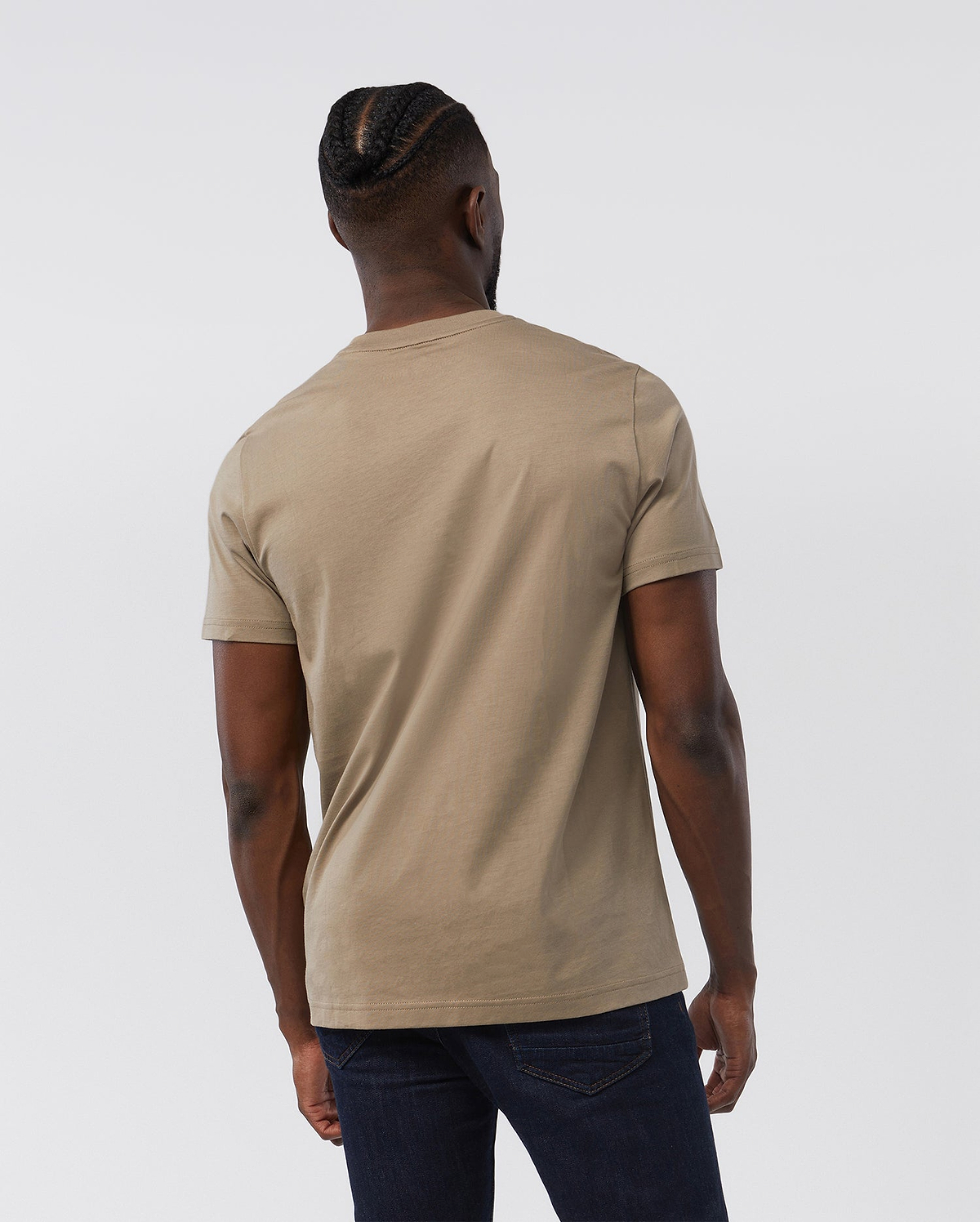A person with cornrowed hair is seen from the back, wearing the Psycho Bunny MENS CLASSIC CREW NECK TEE - B6U014CRPC made of 100% Pima cotton and dark jeans. The light beige short-sleeve shirt serves as the basis for any man's wardrobe, standing out against the plain white background and highlighting the individual's casual attire and hairstyle.