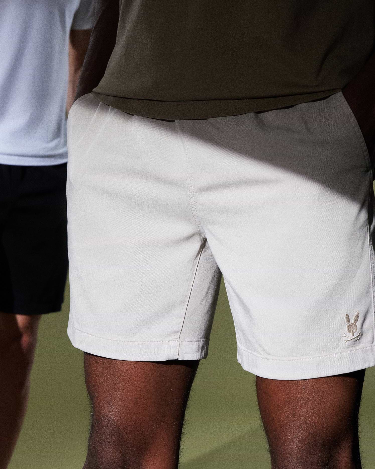 Two people are partially visible. The person in the foreground is wearing light beige shorts, reminiscent of the MENS WILLIS STRETCH TENCEL SHORT by Psycho Bunny, while the person in the background sports black shorts and a white shirt. The photo spotlights the shorts against a plain background.