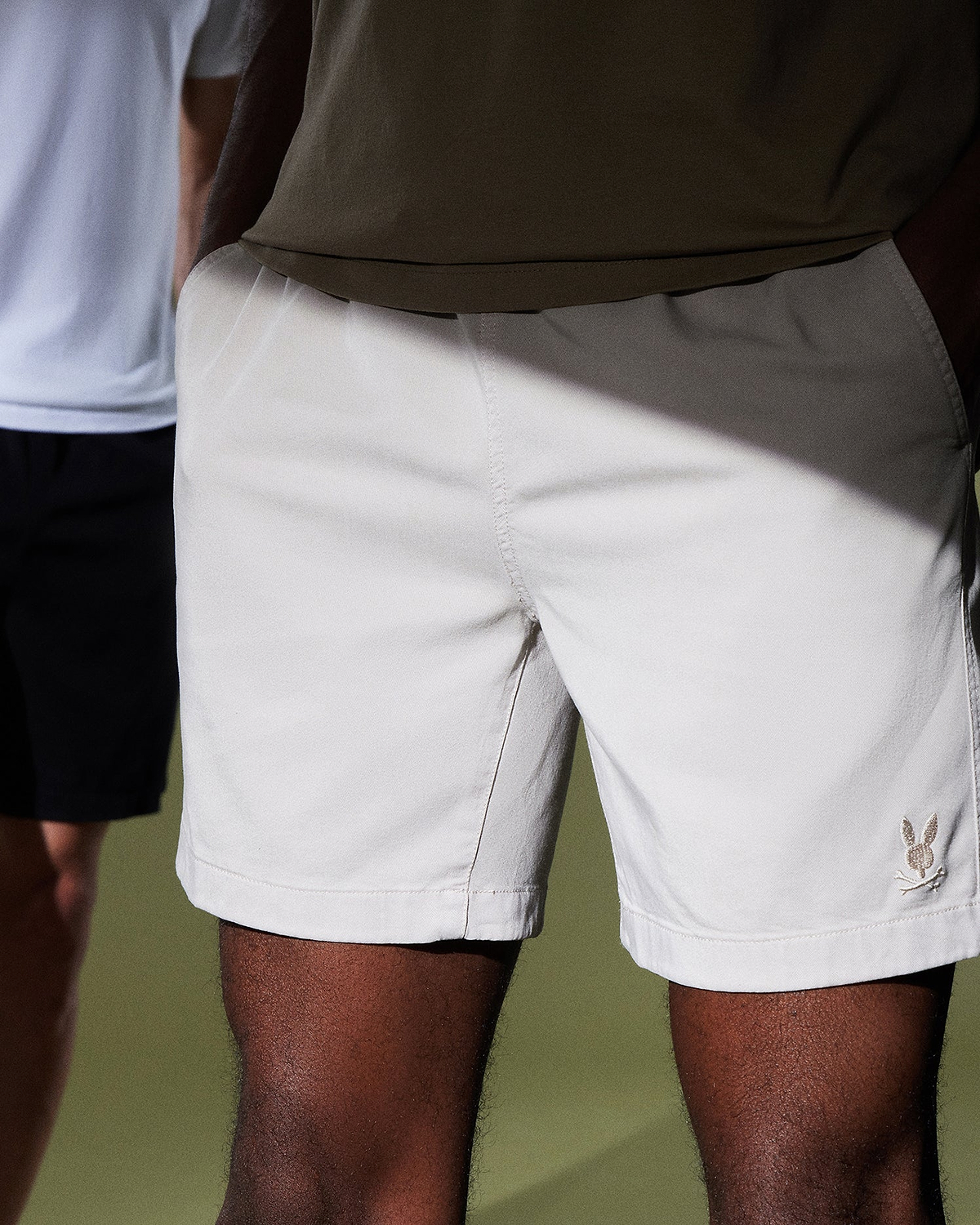 Two people are partially visible. The person in the foreground is wearing light beige shorts, reminiscent of the MENS WILLIS STRETCH TENCEL SHORT by Psycho Bunny, while the person in the background sports black shorts and a white shirt. The photo spotlights the shorts against a plain background.