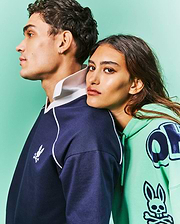 Navy blue classic fit rugby shirt with white collar, piping, and small embroidered Psycho Bunny logo. Mint green Mens Toby Oversized Hoodie featuring bold dark blue graphic letters and large Psycho Bunny skull print.