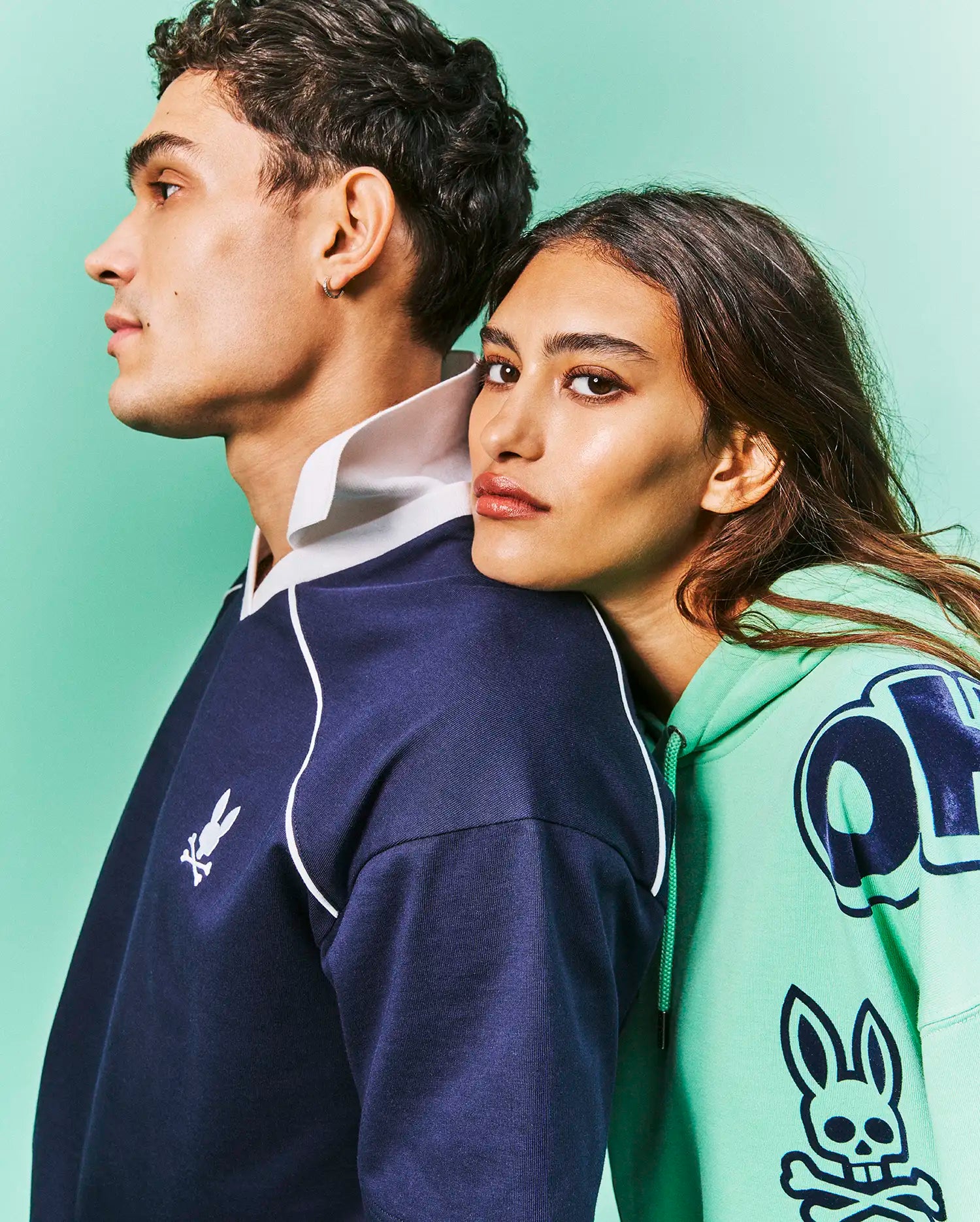 Navy blue classic fit rugby shirt with white collar, piping, and small embroidered Psycho Bunny logo. Mint green Mens Toby Oversized Hoodie featuring bold dark blue graphic letters and large Psycho Bunny skull print.