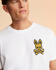 A person wearing a white Psycho Bunny MENS VEGAS CITY TEE - B6U967X1PC with a distinctive printed Bunny Logo featuring a cartoonish rabbit skull and crossbones on the upper left side is shown. The background is a gradient of warm, peachy tones. The person has short hair and is looking off to the side.