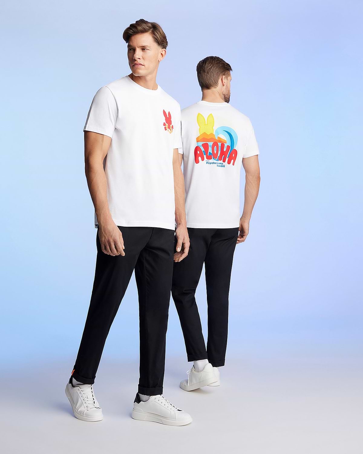 Two men are standing against a light blue background, wearing matching white t-shirts and black pants. The man on the left faces the camera, showcasing a red graphic pattern on his Pima cotton jersey. The man on the right is turned away, revealing a colorful "ALOHA" Hawaii tee design on the back of his MENS HAWAII CITY TEE - B6U169Y1PC by Psycho Bunny.