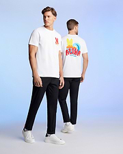 Two men are standing against a light blue background, wearing matching white t-shirts and black pants. The man on the left faces the camera, showcasing a red graphic pattern on his Pima cotton jersey. The man on the right is turned away, revealing a colorful "ALOHA" Hawaii tee design on the back of his MENS HAWAII CITY TEE - B6U169Y1PC by Psycho Bunny.