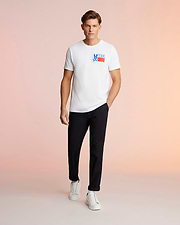 A man stands against a pink background, wearing a white Psycho Bunny MENS TEXAS TEE - B6U553W1PC with a "Deus Ex Machina" logo, black trousers, and white sneakers, looking directly at the camera. This T-shirt is made from Pima cotton jersey.