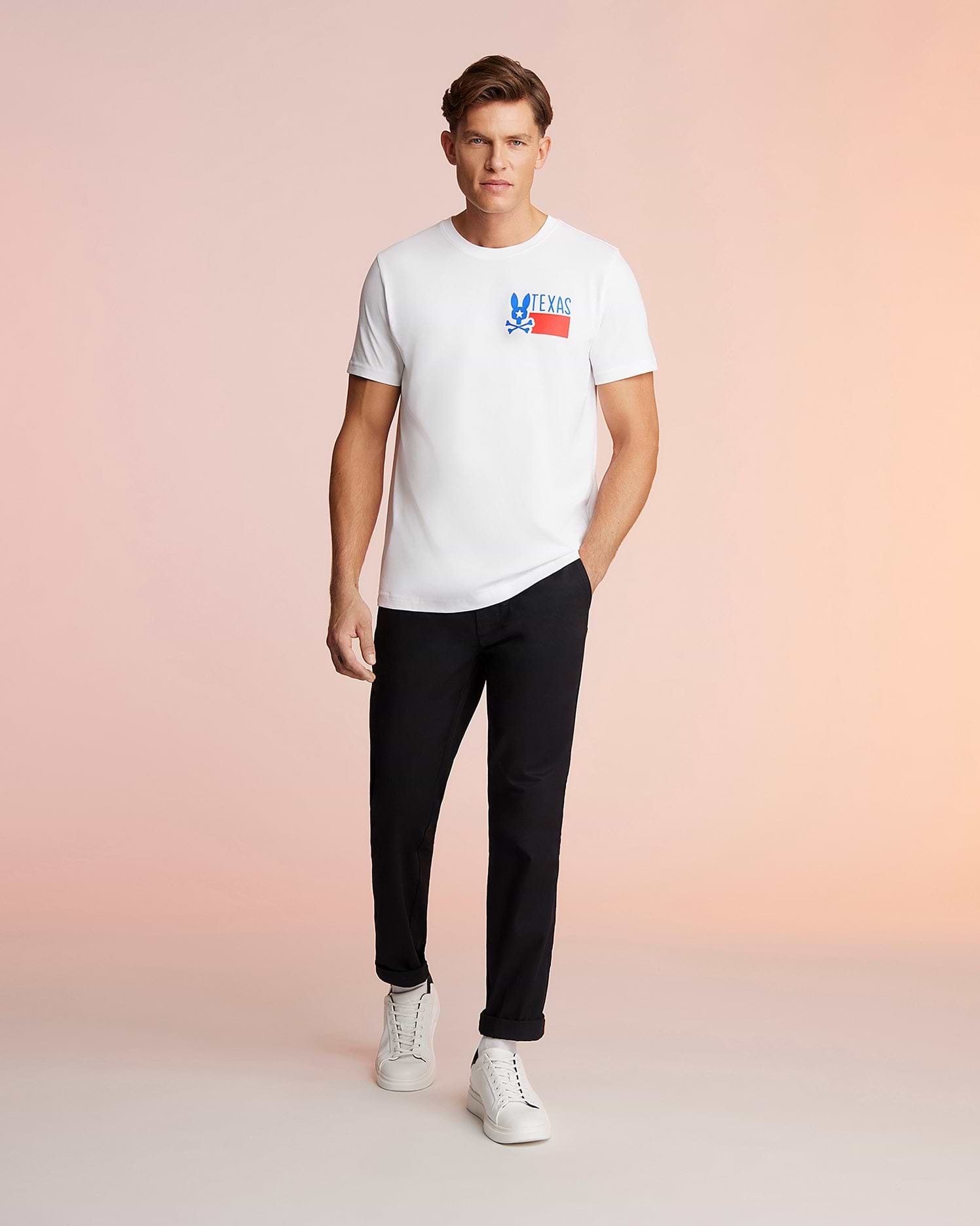 A man stands against a pink background, wearing a white Psycho Bunny MENS TEXAS TEE - B6U553W1PC with a "Deus Ex Machina" logo, black trousers, and white sneakers, looking directly at the camera. This T-shirt is made from Pima cotton jersey.