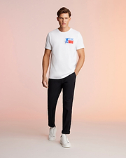 A man stands against a pink background, wearing a white Psycho Bunny MENS TEXAS TEE - B6U553W1PC with a "Deus Ex Machina" logo, black trousers, and white sneakers, looking directly at the camera. This T-shirt is made from Pima cotton jersey.