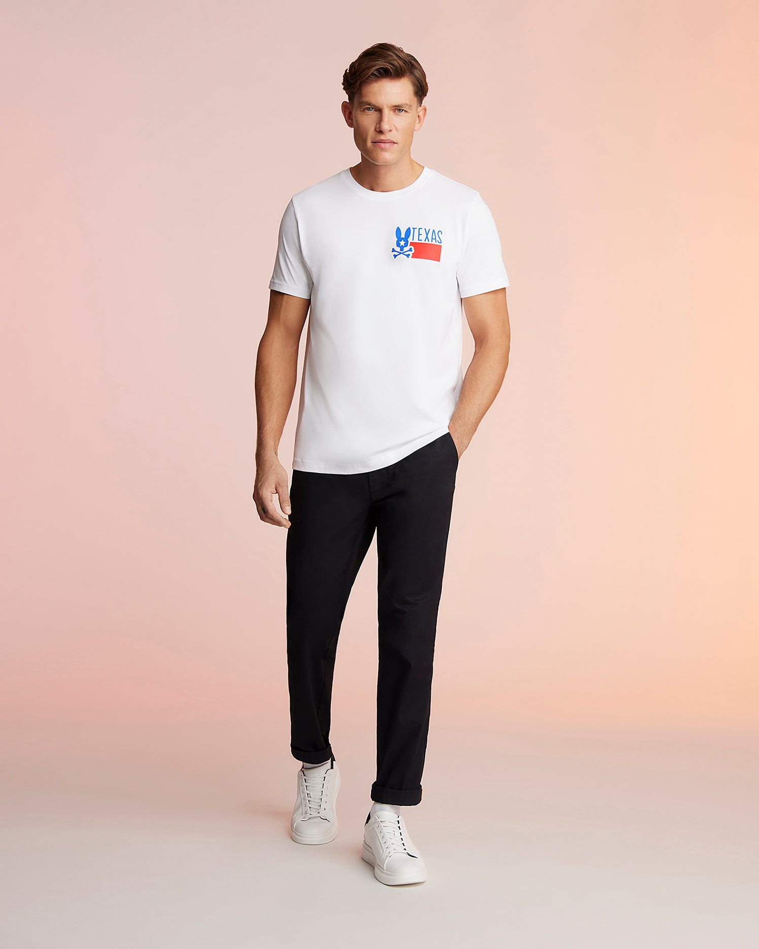 A man stands against a pink background, wearing a white Psycho Bunny MENS TEXAS TEE - B6U553W1PC with a "Deus Ex Machina" logo, black trousers, and white sneakers, looking directly at the camera. This T-shirt is made from Pima cotton jersey.