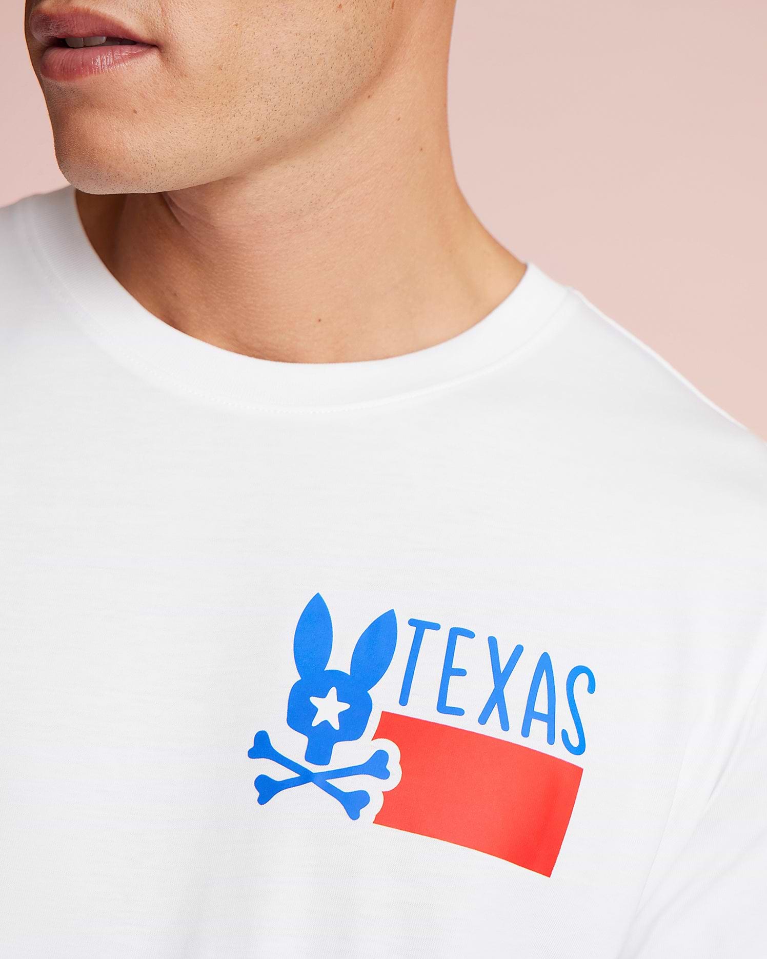 Close-up of a man wearing a white Psycho Bunny Pima cotton jersey T-shirt with a blue and red graphic of a stylized rabbit over the word "TEXAS." The background is a soft pink.