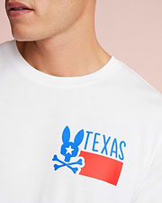 Close-up of a man wearing a white Psycho Bunny Pima cotton jersey T-shirt with a blue and red graphic of a stylized rabbit over the word "TEXAS." The background is a soft pink.